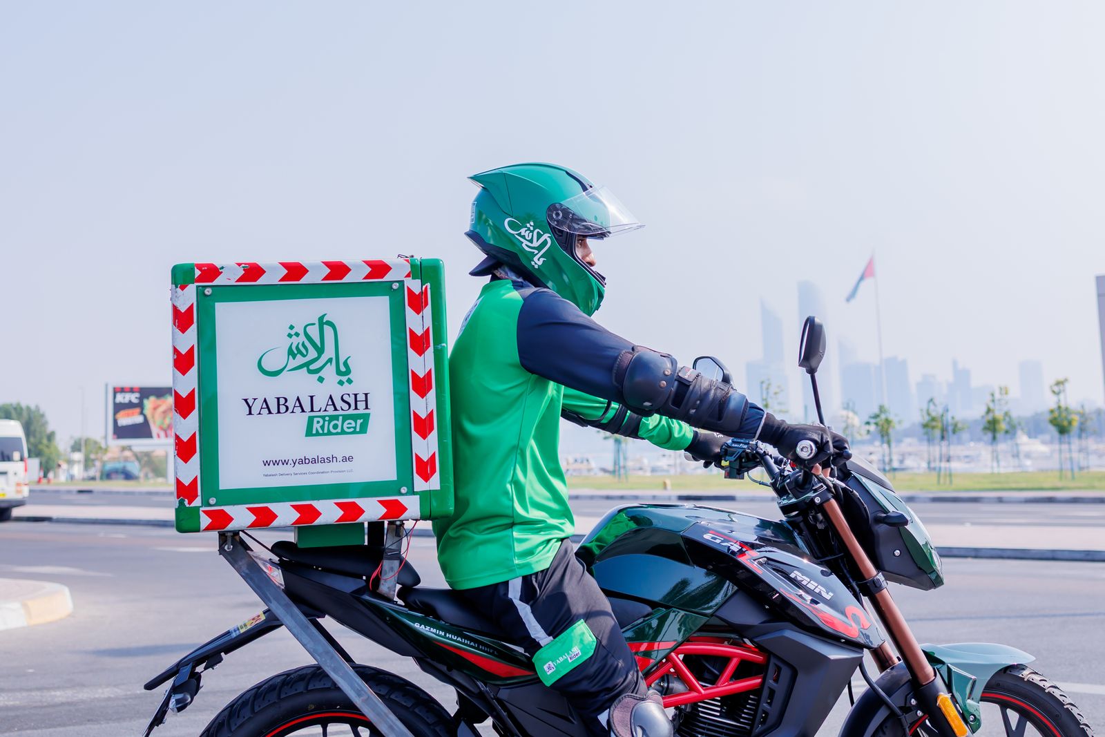 A YaBalash delivery rider on a branded motorcycle with the Abu Dhabi skyline behind.